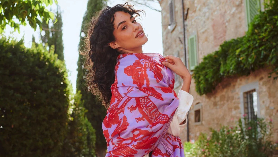 Woman with a colorful scarf standing in a sunlit street with greenery and a stone building.