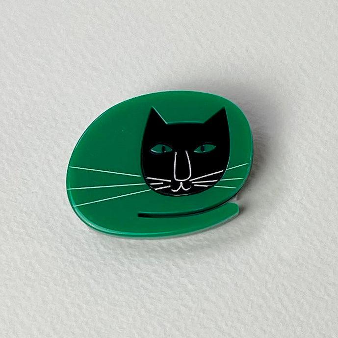 Sleek glossy green enamel pin of stylized black cat with teal eyes and white whiskers